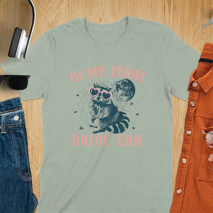 A raccoon wearing heart-shaped sunglasses and a bride's veil, with the moon in the background. Text reads In My Feral Bride Era.