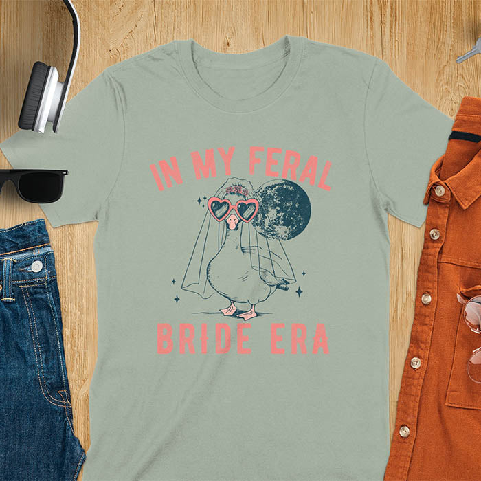 A goose standing, wearing heart-shaped sunglasses and a bride's veil. Moon in the upper right background. Retro-style text reads In My Feral on top Bride Era at bottom. Whimsical image.