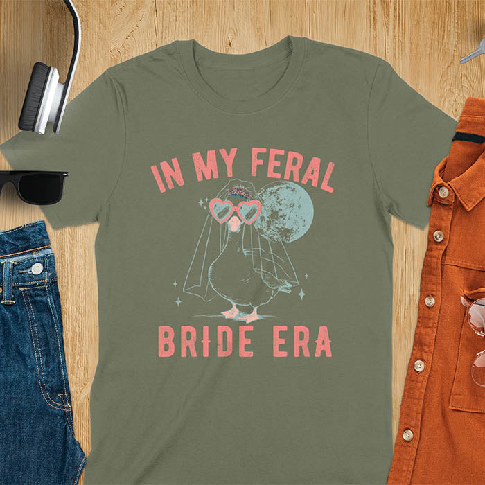 A goose standing, wearing heart-shaped sunglasses and a bride's veil. Moon in the upper right background. Retro-style text reads In My Feral on top Bride Era at bottom. Whimsical image.