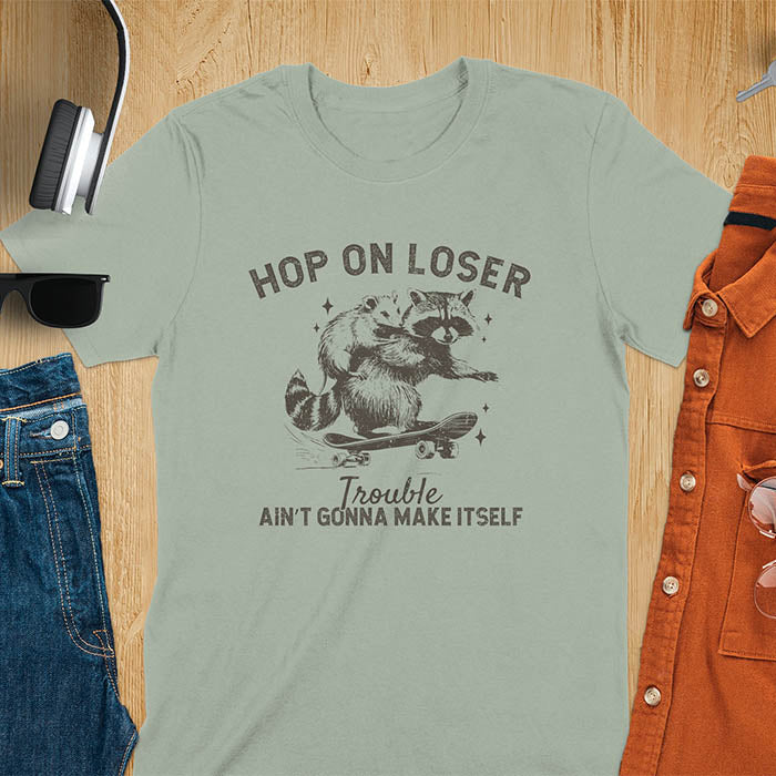 A raccoon riding a skateboard with an opossum on its back. Text reads Hop On Loser and Trouble Ain't Gonna Make Itself.