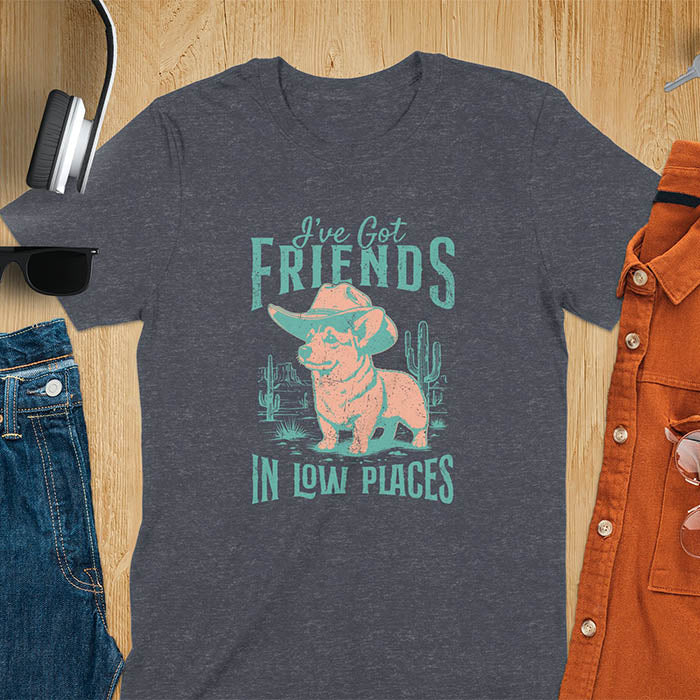 A t-shirt with a corgi wearing a cowboy hat standing in a desert scene with cacti. Text: "I've Got FRIENDS" above and "IN LOW PLACES" below. This retro dog shirt is a corgi shirt and funny dog shirt, a vintage dog tee. A cowboy tshirt for a dog dad shirt and pet owner gift. An animal person gift with cuteness puppy, a meme graphic tee, a mom gift option.