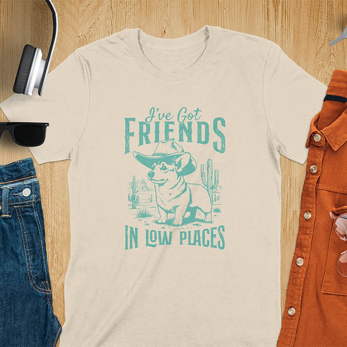 A t-shirt with a corgi wearing a cowboy hat standing in a desert scene with cacti. Text: "I've Got FRIENDS" above and "IN LOW PLACES" below. This retro dog shirt is a corgi shirt and funny dog shirt, a vintage dog tee. A cowboy tshirt for a dog dad shirt and pet owner gift. An animal person gift with cuteness puppy, a meme graphic tee, a mom gift option.