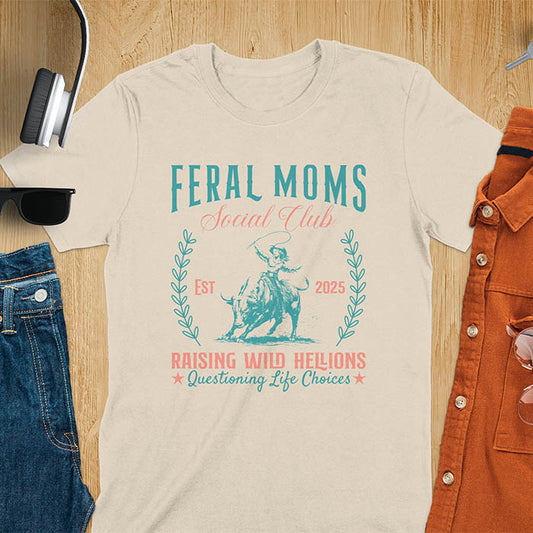 A t-shirt with a cowgirl riding a bucking bull. Text: Feral Moms SOCIAL CLUB above and Raising HELLIONS, questioning life choices below. This unhinged t-shirts design features a cowgirl mom on a rodeo bucking bull, a weird meme shirt for a feral mom gift. A retro inspired design, one of those shirts that go hard, a mama outfit or feral wife gift. A mom gift or gift for new mom for a western mama.