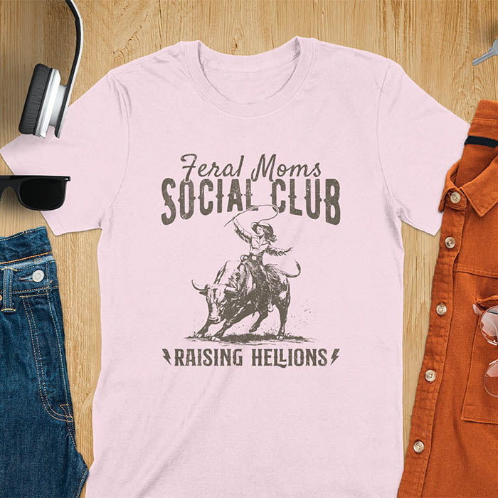 A t-shirt with a cowgirl riding a bucking bull. Text: Feral Moms SOCIAL CLUB above and Raising HELLIONS below. This unhinged t-shirts design features a cowgirl mom on a rodeo bucking bull, a weird meme shirt for a feral mom gift. A retro inspired design, one of those shirts that go hard, a mama outfit or feral wife gift. A mom gift or gift for new mom for a western mama.