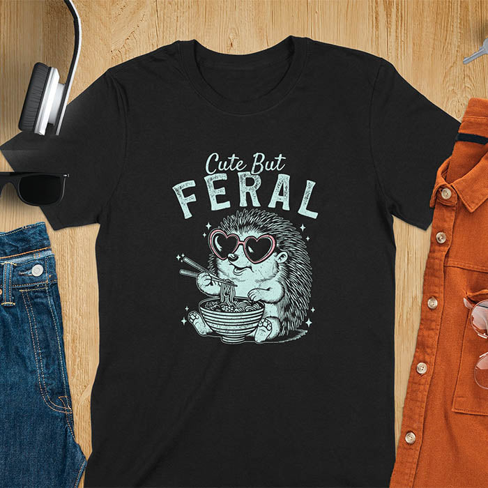 A t-shirt with a hedgehog wearing heart sunglasses and eating ramen with chopsticks. Text: "Cute But" above and "FERAL" below. This kawaiiness cuteness shows hedgehog cuteness in a weird funny shirt while ramen eating, a funny animal meme. A meme graphic tee and funny unhinge shirt, a great animal person gift and pet owner gift. A gen z meme with retro aesthetics tee vibes, a gift for mom or hers option, not quite wild safari animals.