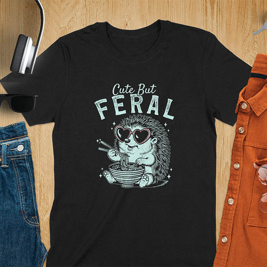 A t-shirt with a hedgehog wearing heart sunglasses and eating ramen with chopsticks. Text: "Cute But" above and "FERAL" below. This kawaiiness cuteness shows hedgehog cuteness in a weird funny shirt while ramen eating, a funny animal meme. A meme graphic tee and funny unhinge shirt, a great animal person gift and pet owner gift. A gen z meme with retro aesthetics tee vibes, a gift for mom or hers option, not quite wild safari animals.