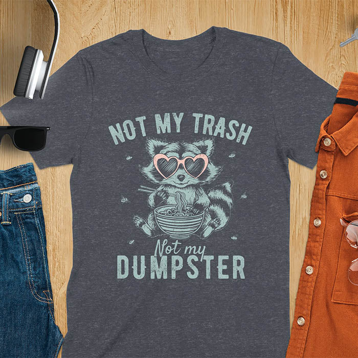 A t-shirt featuring a raccoon wearing pink heart-shaped sunglasses, sitting with chopsticks and eating ramen from a bowl. Small flies surround the raccoon. Text: NOT MY TRASH above and Not my DUMPSTER below in a distressed, bold font. The raccoon has a striped tail and a cartoonish appearance.