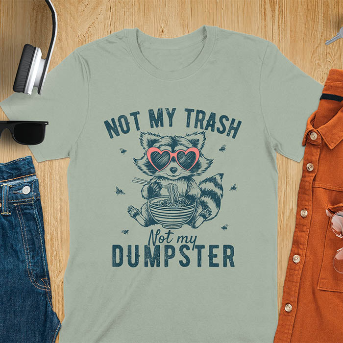 A t-shirt featuring a raccoon wearing pink heart-shaped sunglasses, sitting with chopsticks and eating ramen from a bowl. Small flies surround the raccoon. Text: NOT MY TRASH above and Not my DUMPSTER below in a distressed, bold font. The raccoon has a striped tail and a cartoonish appearance.