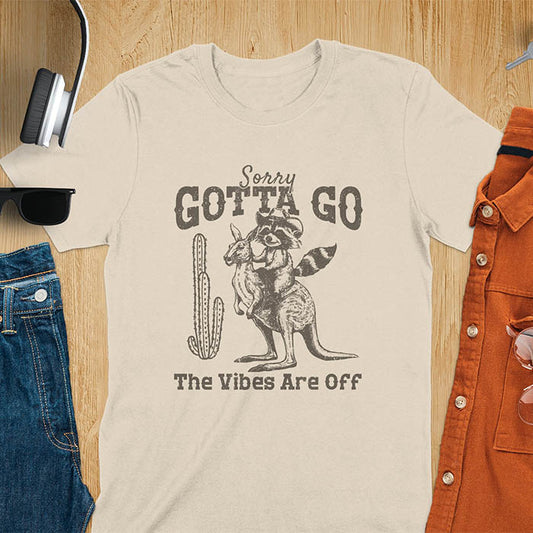 A t-shirt with a raccoon wearing a cowboy hat riding a kangaroo. Text: Sorry GOTTA GO above and The Vibes Are Off below. This humorous tee-shirt features a funny gen z meme and sarcastic meme shirt, a weird graphics shirt.  A funny retro graphic and vintage graphic tee with rodeo cowboy elements. A silly shirt or funny unhinged shirt, an animal person gift or kangaroo gifts. A raccoon t shirt or mom gift alternative.