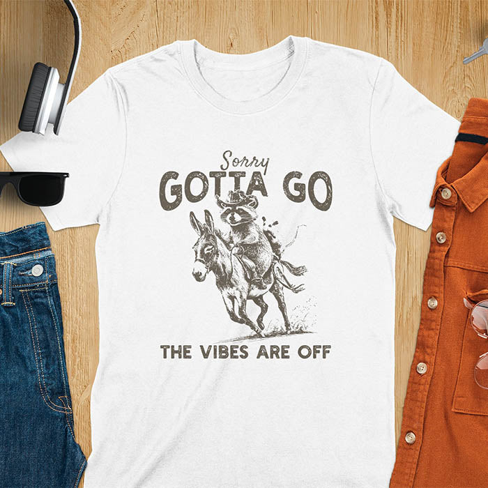 A t-shirt with a raccoon wearing a cowboy hat riding a donkey, with an opossum seated behind. Text: Sorry GOTTA GO above and THE VIBES ARE OFF below. This humorous T-shirt features a funny donkey and opossum possum, a weird graphics shirt. A sarcastic meme shirt and funny Gen Z meme, with rodeo cowboy elements. This vintage graphic is one of those shirts that go hard, an animal person gift or raccoon person gift. A funny unhinged shirt or mom gift alternative.