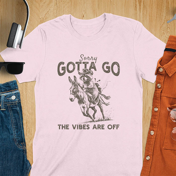 A t-shirt with a raccoon wearing a cowboy hat riding a donkey, with an opossum seated behind. Text: Sorry GOTTA GO above and THE VIBES ARE OFF below. This humorous T-shirt features a funny donkey and opossum possum, a weird graphics shirt. A sarcastic meme shirt and funny Gen Z meme, with rodeo cowboy elements. This vintage graphic is one of those shirts that go hard, an animal person gift or raccoon person gift. A funny unhinged shirt or mom gift alternative.