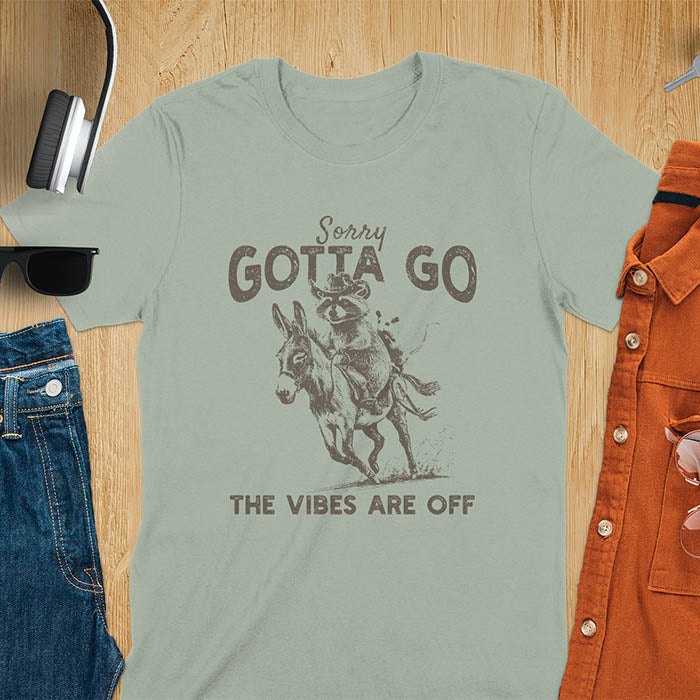 A t-shirt with a raccoon wearing a cowboy hat riding a donkey, with an opossum seated behind. Text: Sorry GOTTA GO above and THE VIBES ARE OFF below. This humorous T-shirt features a funny donkey and opossum possum, a weird graphics shirt. A sarcastic meme shirt and funny Gen Z meme, with rodeo cowboy elements. This vintage graphic is one of those shirts that go hard, an animal person gift or raccoon person gift. A funny unhinged shirt or mom gift alternative.