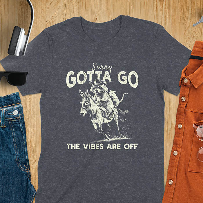 A t-shirt with a raccoon wearing a cowboy hat riding a donkey, with an opossum seated behind. Text: Sorry GOTTA GO above and THE VIBES ARE OFF below. This humorous T-shirt features a funny donkey and opossum possum, a weird graphics shirt. A sarcastic meme shirt and funny Gen Z meme, with rodeo cowboy elements. This vintage graphic is one of those shirts that go hard, an animal person gift or raccoon person gift. A funny unhinged shirt or mom gift alternative.