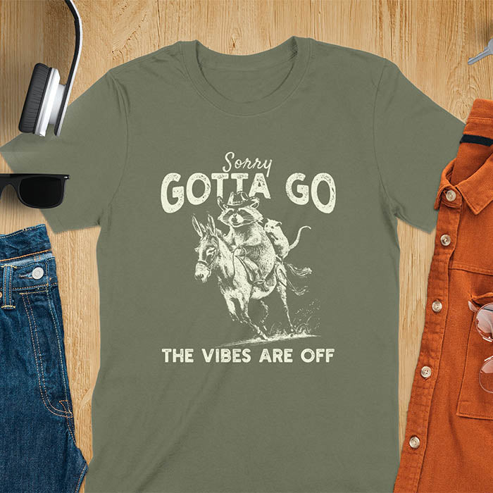 A t-shirt with a raccoon wearing a cowboy hat riding a donkey, with an opossum seated behind. Text: Sorry GOTTA GO above and THE VIBES ARE OFF below. This humorous T-shirt features a funny donkey and opossum possum, a weird graphics shirt. A sarcastic meme shirt and funny Gen Z meme, with rodeo cowboy elements. This vintage graphic is one of those shirts that go hard, an animal person gift or raccoon person gift. A funny unhinged shirt or mom gift alternative.