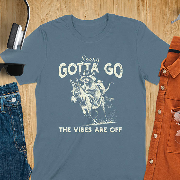 A t-shirt with a raccoon wearing a cowboy hat riding a donkey, with an opossum seated behind. Text: Sorry GOTTA GO above and THE VIBES ARE OFF below. This humorous T-shirt features a funny donkey and opossum possum, a weird graphics shirt. A sarcastic meme shirt and funny Gen Z meme, with rodeo cowboy elements. This vintage graphic is one of those shirts that go hard, an animal person gift or raccoon person gift. A funny unhinged shirt or mom gift alternative.