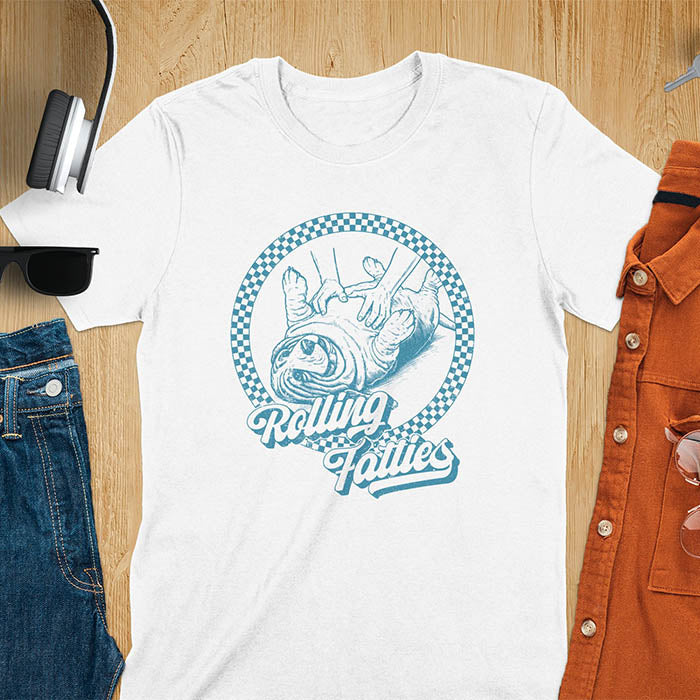 A T-shirt design featuring a detailed illustration of a chubby basset hound being rolled by two hands, surrounded by a checkered circle. The text Rolling Fatties is written in a stylized font below the image. Retro style with distressed texture