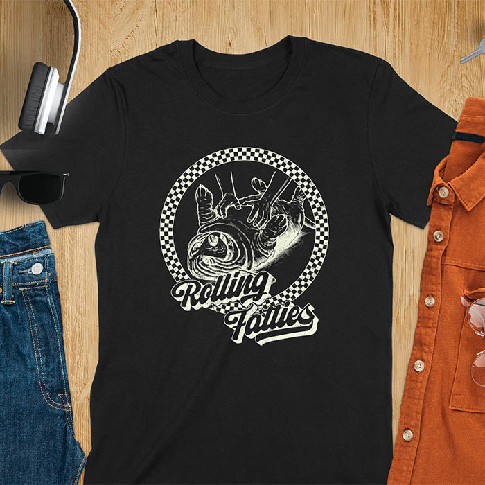 A T-shirt design featuring a detailed illustration of a chubby basset hound being rolled by two hands, surrounded by a checkered circle. The text Rolling Fatties is written in a stylized font below the image. Retro style with distressed texture