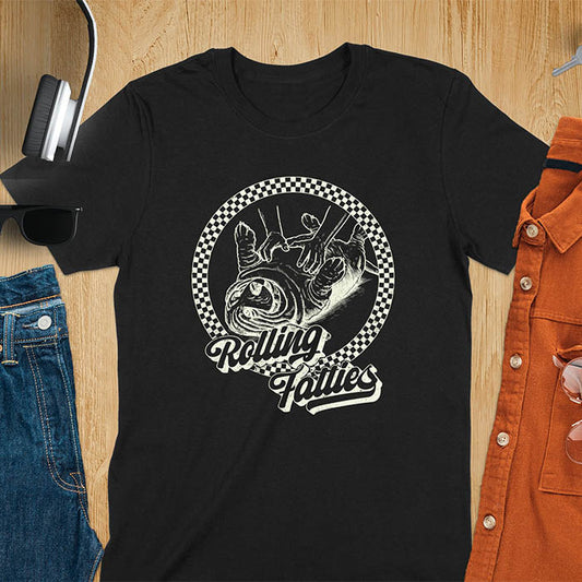 A T-shirt design featuring a detailed illustration of a chubby basset hound being rolled by two hands, surrounded by a checkered circle. The text Rolling Fatties is written in a stylized font below the image. Retro style with distressed texture