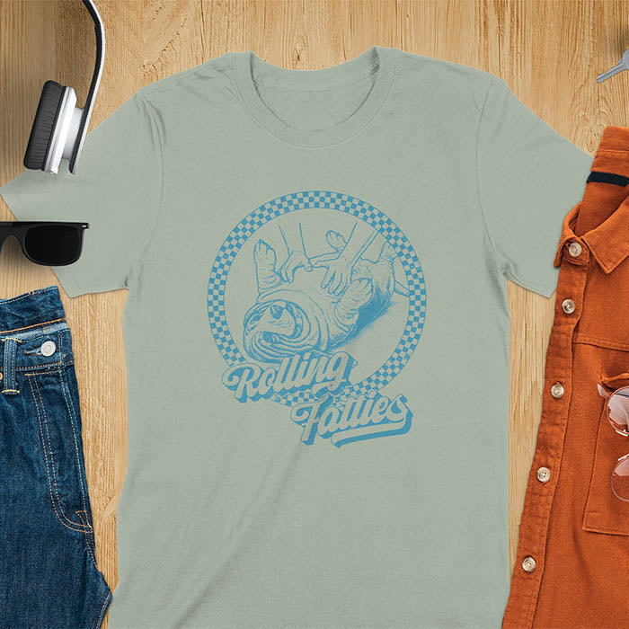 A T-shirt design featuring a detailed illustration of a chubby basset hound being rolled by two hands, surrounded by a checkered circle. The text Rolling Fatties is written in a stylized font below the image. Retro style with distressed texture