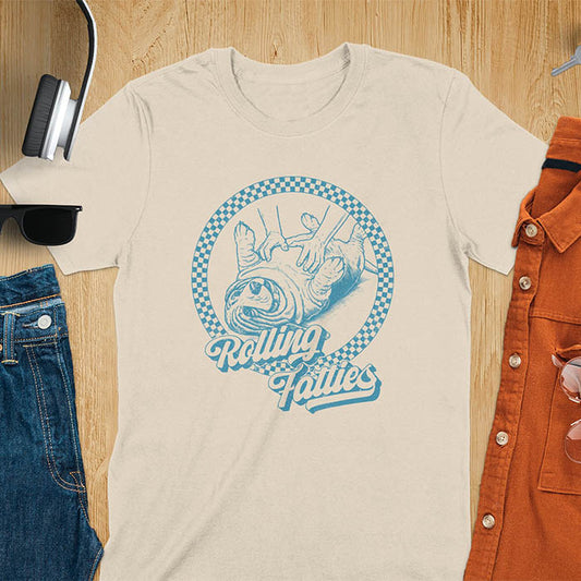 A T-shirt design featuring a detailed illustration of a chubby basset hound being rolled by two hands, surrounded by a checkered circle. The text Rolling Fatties is written in a stylized font below the image. Retro style with distressed texture