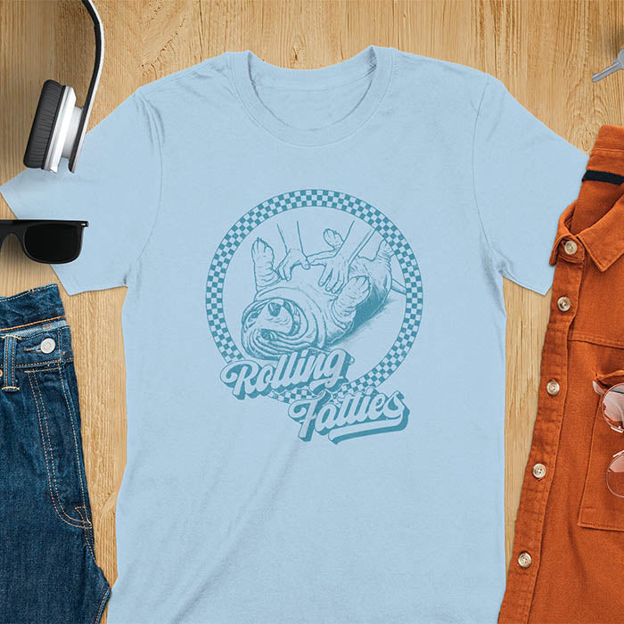 A T-shirt design featuring a detailed illustration of a chubby basset hound being rolled by two hands, surrounded by a checkered circle. The text Rolling Fatties is written in a stylized font below the image. Retro style with distressed texture