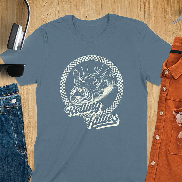 A T-shirt design featuring a detailed illustration of a chubby basset hound being rolled by two hands, surrounded by a checkered circle. The text Rolling Fatties is written in a stylized font below the image. Retro style with distressed texture