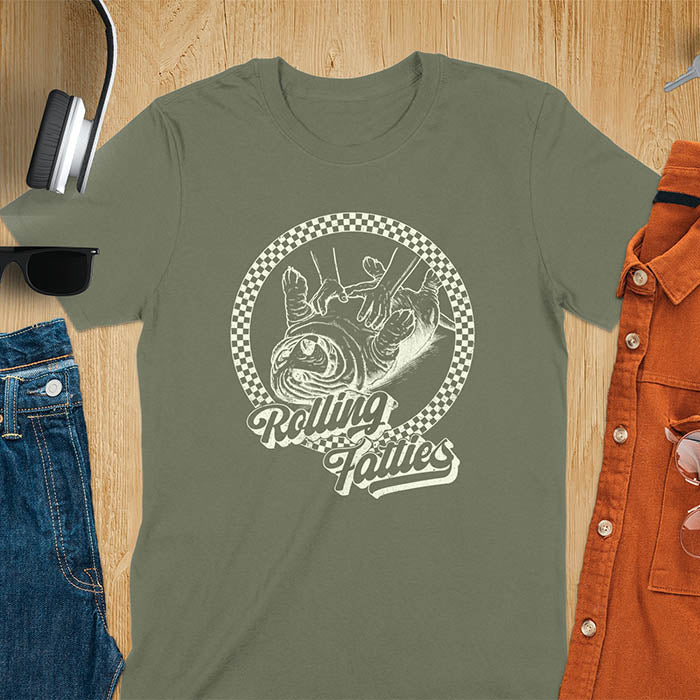 A T-shirt design featuring a detailed illustration of a chubby basset hound being rolled by two hands, surrounded by a checkered circle. The text Rolling Fatties is written in a stylized font below the image. Retro style with distressed texture