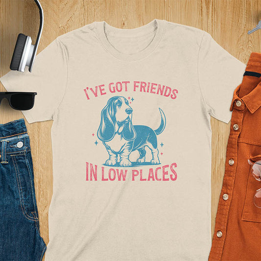 This retro dog shirt features a vintage dog tee design of a basset hound standing proudly, tail high, and gazing to the side. Text reads “I’ve got friends” above and “in low places” below. A distressed texture adds charm to the shirt.