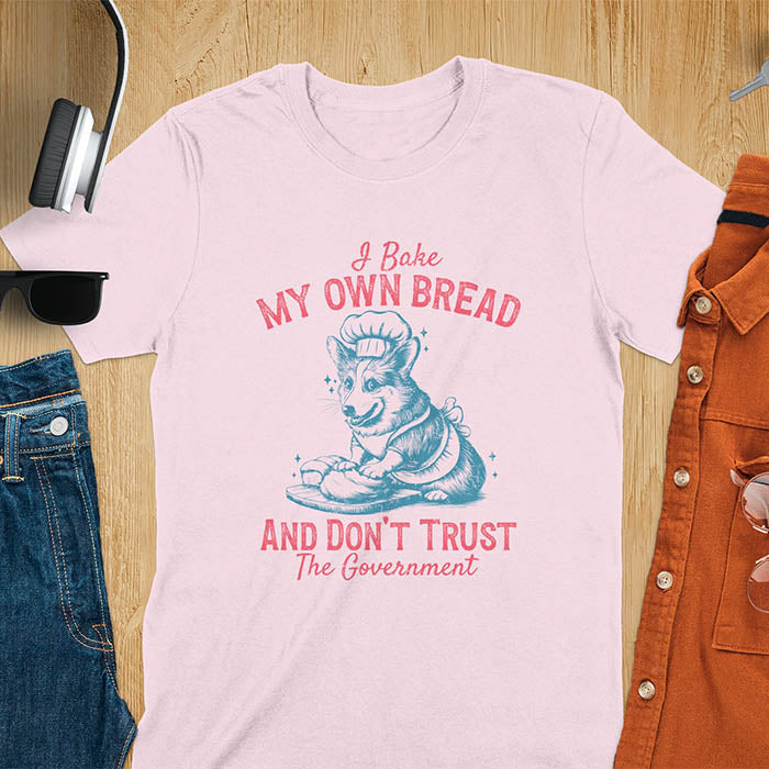 A t-shirt with a corgi wearing a chef's hat and apron, kneading dough. Text: "I Bake MY OWN BREAD" above and "AND DON'T TRUST The Government" below. This retro sarcastic tee is a funny meme shirt and funny dog shirt, a corgi shirt for a corgi lovers gift. An animal lovers tshirt and dog lover gift, a dog mom shirt or pet dad shirt. A dog person tee and pet dog owner gift, one of the funny bakers gifts, a gift for mom or her.