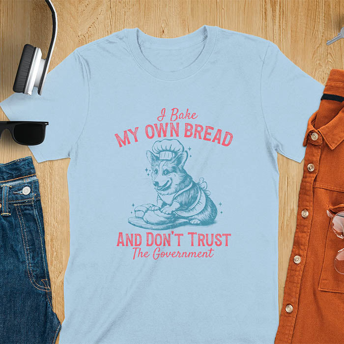 A t-shirt with a corgi wearing a chef's hat and apron, kneading dough. Text: "I Bake MY OWN BREAD" above and "AND DON'T TRUST The Government" below. This retro sarcastic tee is a funny meme shirt and funny dog shirt, a corgi shirt for a corgi lovers gift. An animal lovers tshirt and dog lover gift, a dog mom shirt or pet dad shirt. A dog person tee and pet dog owner gift, one of the funny bakers gifts, a gift for mom or her.