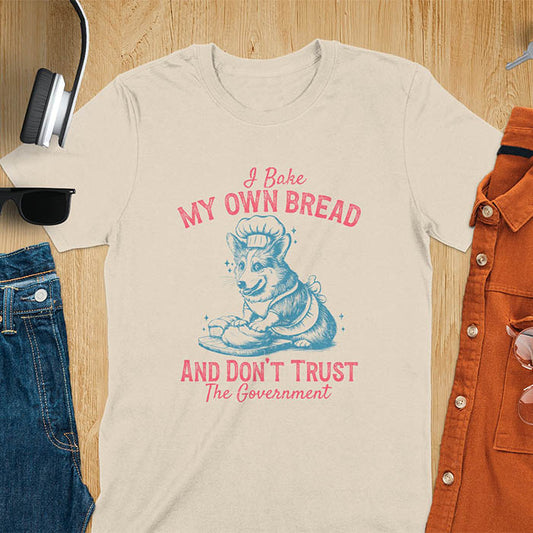 A t-shirt with a corgi wearing a chef's hat and apron, kneading dough. Text: "I Bake MY OWN BREAD" above and "AND DON'T TRUST The Government" below. This retro sarcastic tee is a funny meme shirt and funny dog shirt, a corgi shirt for a corgi lovers gift. An animal lovers tshirt and dog lover gift, a dog mom shirt or pet dad shirt. A dog person tee and pet dog owner gift, one of the funny bakers gifts, a gift for mom or her.