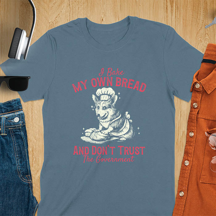 A t-shirt with a corgi wearing a chef's hat and apron, kneading dough. Text: "I Bake MY OWN BREAD" above and "AND DON'T TRUST The Government" below. This retro sarcastic tee is a funny meme shirt and funny dog shirt, a corgi shirt for a corgi lovers gift. An animal lovers tshirt and dog lover gift, a dog mom shirt or pet dad shirt. A dog person tee and pet dog owner gift, one of the funny bakers gifts, a gift for mom or her.