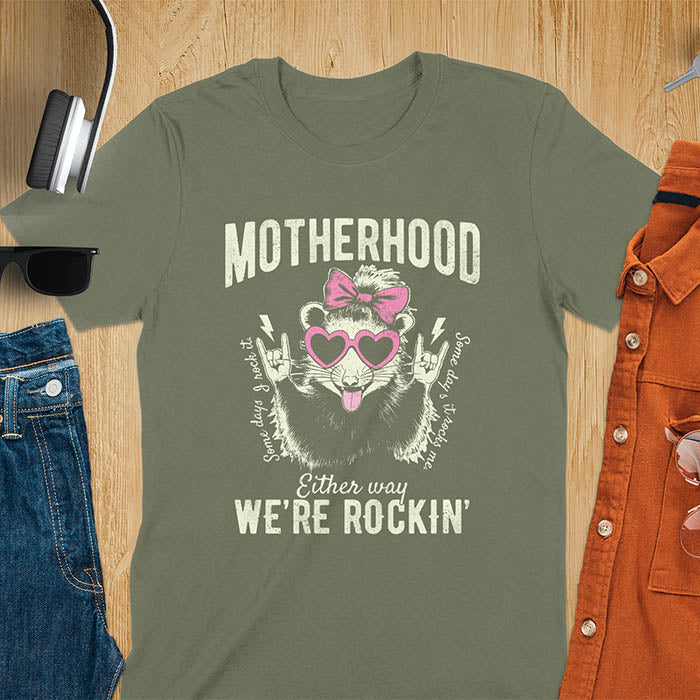 A t-shirt with an opossum wearing a bow and heart-shaped sunglasses, throwing rock-on hand gestures. Text: MOTHERHOOD above, Some days I rock it left, Some days it rocks me right, and Either way WE'RE ROCKIN below. Lightning bolt accents on the hands. This vintage mama tee or rocker mom shirt is a trending mama design with a mom saying. A snarky humor designs and unhinged shirts style, a mom gift hers or baby shower gift. A first time mom gift or part of mommy outfits, for rocking motherhood.