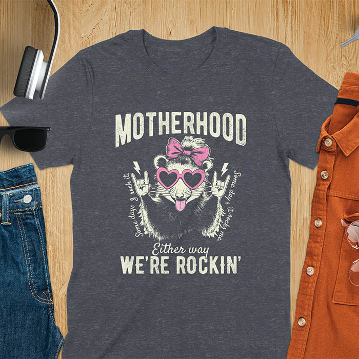 A t-shirt with an opossum wearing a bow and heart-shaped sunglasses, throwing rock-on hand gestures. Text: MOTHERHOOD above, Some days I rock it left, Some days it rocks me right, and Either way WE'RE ROCKIN below. Lightning bolt accents on the hands. This vintage mama tee or rocker mom shirt is a trending mama design with a mom saying. A snarky humor designs and unhinged shirts style, a mom gift hers or baby shower gift. A first time mom gift or part of mommy outfits, for rocking motherhood.