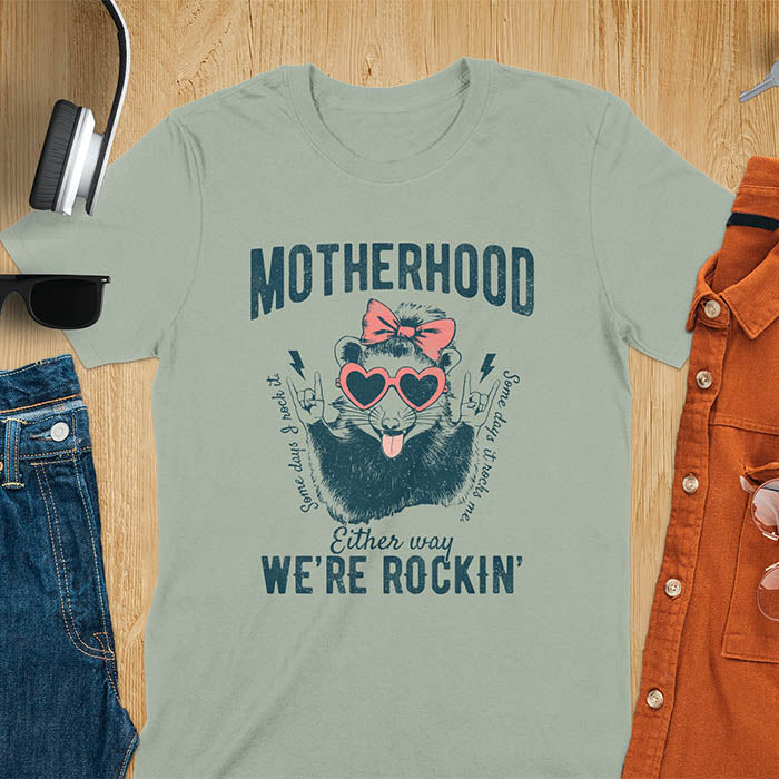 A t-shirt with an opossum wearing a bow and heart-shaped sunglasses, throwing rock-on hand gestures. Text: MOTHERHOOD above, Some days I rock it left, Some days it rocks me right, and Either way WE'RE ROCKIN below. Lightning bolt accents on the hands. This vintage mama tee or rocker mom shirt is a trending mama design with a mom saying. A snarky humor designs and unhinged shirts style, a mom gift hers or baby shower gift. A first time mom gift or part of mommy outfits, for rocking motherhood.