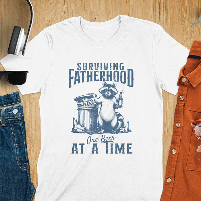 A t-shirt with a raccoon holding a beer next to a overflowing trash can. Text: "SURVIVING" above "FATHERHOOD" and "One Beer" above "AT A TIME" below. This Fatherhood shirt is a dad t shirt and dad humor gift. A Fathers day gift or Fathers day shirt, a dad birthday shirt and dad gift. A Funny father shirt and gift for daddie, a new daddy gift. Part of a husband outfit and Funny dad clothe, for surviving fatherhood.