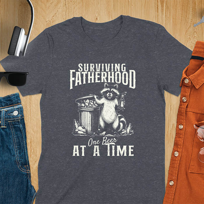 A t-shirt with a raccoon holding a beer next to a overflowing trash can. Text: "SURVIVING" above "FATHERHOOD" and "One Beer" above "AT A TIME" below. This Fatherhood shirt is a dad t shirt and dad humor gift. A Fathers day gift or Fathers day shirt, a dad birthday shirt and dad gift. A Funny father shirt and gift for daddie, a new daddy gift. Part of a husband outfit and Funny dad clothe, for surviving fatherhood.