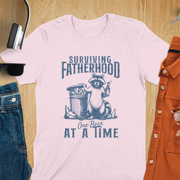 A t-shirt with a raccoon holding a beer next to a overflowing trash can. Text: "SURVIVING" above "FATHERHOOD" and "One Beer" above "AT A TIME" below. This Fatherhood shirt is a dad t shirt and dad humor gift. A Fathers day gift or Fathers day shirt, a dad birthday shirt and dad gift. A Funny father shirt and gift for daddie, a new daddy gift. Part of a husband outfit and Funny dad clothe, for surviving fatherhood.