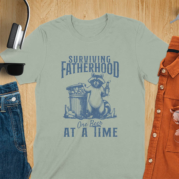 A t-shirt with a raccoon holding a beer next to a overflowing trash can. Text: "SURVIVING" above "FATHERHOOD" and "One Beer" above "AT A TIME" below. This Fatherhood shirt is a dad t shirt and dad humor gift. A Fathers day gift or Fathers day shirt, a dad birthday shirt and dad gift. A Funny father shirt and gift for daddie, a new daddy gift. Part of a husband outfit and Funny dad clothe, for surviving fatherhood.