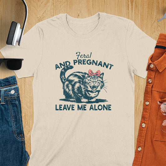 A t-shirt with a tabby cat with a grumpy expression and a pink bow. Text: Feral AND PREGNANT above and LEAVE ME ALONE below. This funny maternity tee is a feral cats shirt for a mom to be shirt. A pregnancy humor design for a pregnancy announce or sibling pregnancy. An expecting mom gift or mommy to be gift, a maternity apparel and pregnancy keepsake for an expecting baby during a feral pregnancy.