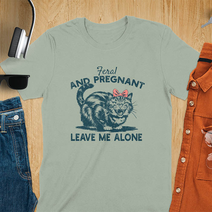 A t-shirt with a tabby cat with a grumpy expression and a pink bow. Text: Feral AND PREGNANT above and LEAVE ME ALONE below. This funny maternity tee is a feral cats shirt for a mom to be shirt. A pregnancy humor design for a pregnancy announce or sibling pregnancy. An expecting mom gift or mommy to be gift, a maternity apparel and pregnancy keepsake for an expecting baby during a feral pregnancy.