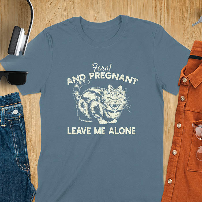 A t-shirt with a tabby cat with a grumpy expression and a pink bow. Text: Feral AND PREGNANT above and LEAVE ME ALONE below. This funny maternity tee is a feral cats shirt for a mom to be shirt. A pregnancy humor design for a pregnancy announce or sibling pregnancy. An expecting mom gift or mommy to be gift, a maternity apparel and pregnancy keepsake for an expecting baby during a feral pregnancy.