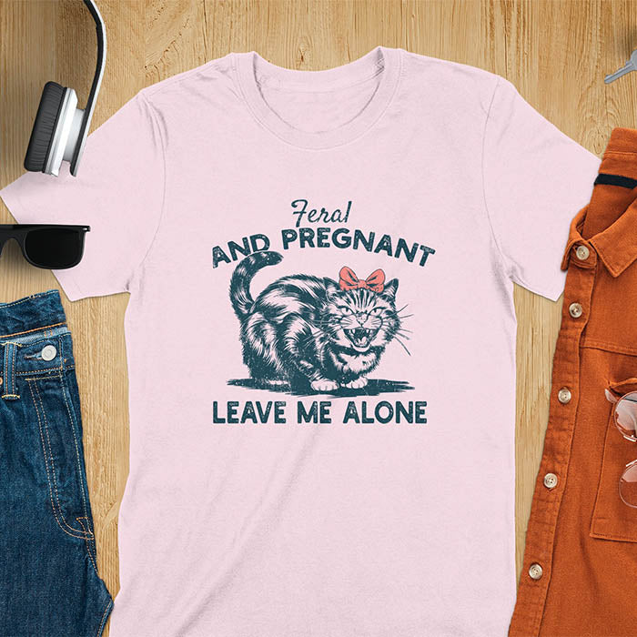 A t-shirt with a tabby cat with a grumpy expression and a pink bow. Text: Feral AND PREGNANT above and LEAVE ME ALONE below. This funny maternity tee is a feral cats shirt for a mom to be shirt. A pregnancy humor design for a pregnancy announce or sibling pregnancy. An expecting mom gift or mommy to be gift, a maternity apparel and pregnancy keepsake for an expecting baby during a feral pregnancy.