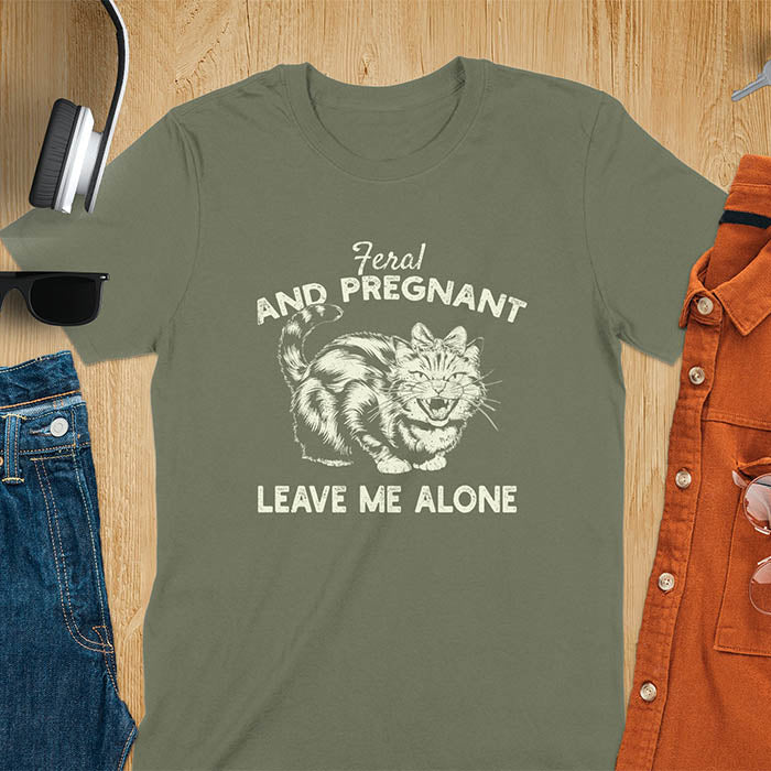 A t-shirt with a tabby cat with a grumpy expression and a pink bow. Text: Feral AND PREGNANT above and LEAVE ME ALONE below. This funny maternity tee is a feral cats shirt for a mom to be shirt. A pregnancy humor design for a pregnancy announce or sibling pregnancy. An expecting mom gift or mommy to be gift, a maternity apparel and pregnancy keepsake for an expecting baby during a feral pregnancy.
