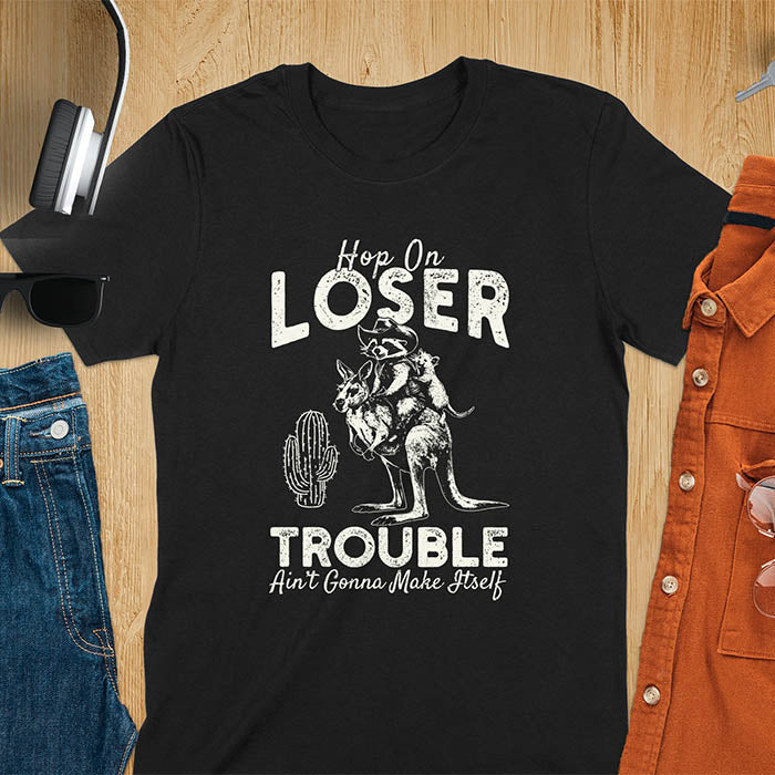 A t-shirt with a raccoon in cowboy attire riding a kangaroo, with an opossum behind. Text: "Hop On LOSER" above and "TROUBLE Ain't Gonna Make Itself" below. This meme t shirt features a funny raccoon (a cute trash panda) dressed as wild west cowboys riding with an opossum possum. A weird graphics shirt with a retro aesthetic, a sarcastic meme shirt and funny genz meme. An animal person gift for a cuteness lover, tapping into weirdcore memes. A mom Gift alternative.
