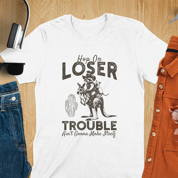 A t-shirt with a raccoon in cowboy attire riding a kangaroo, with an opossum behind. Text: "Hop On LOSER" above and "TROUBLE Ain't Gonna Make Itself" below. This meme t shirt features a funny raccoon (a cute trash panda) dressed as wild west cowboys riding with an opossum possum. A weird graphics shirt with a retro aesthetic, a sarcastic meme shirt and funny genz meme. An animal person gift for a cuteness lover, tapping into weirdcore memes. A mom Gift alternative.