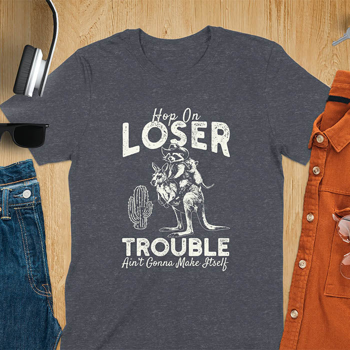 A t-shirt with a raccoon in cowboy attire riding a kangaroo, with an opossum behind. Text: "Hop On LOSER" above and "TROUBLE Ain't Gonna Make Itself" below. This meme t shirt features a funny raccoon (a cute trash panda) dressed as wild west cowboys riding with an opossum possum. A weird graphics shirt with a retro aesthetic, a sarcastic meme shirt and funny genz meme. An animal person gift for a cuteness lover, tapping into weirdcore memes. A mom Gift alternative.