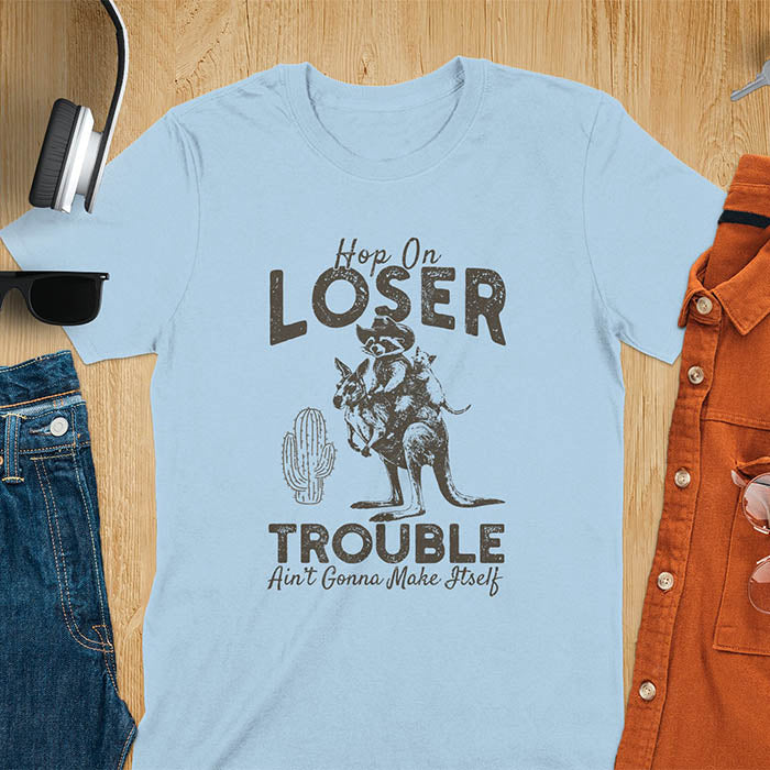 A t-shirt with a raccoon in cowboy attire riding a kangaroo, with an opossum behind. Text: "Hop On LOSER" above and "TROUBLE Ain't Gonna Make Itself" below. This meme t shirt features a funny raccoon (a cute trash panda) dressed as wild west cowboys riding with an opossum possum. A weird graphics shirt with a retro aesthetic, a sarcastic meme shirt and funny genz meme. An animal person gift for a cuteness lover, tapping into weirdcore memes. A mom Gift alternative.