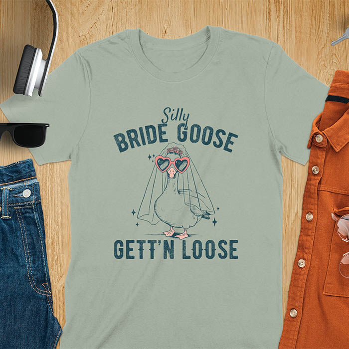 A t-shirt with a goose dressed as a bride with heart sunglasses and a veil. Text: "Silly BRIDE GOOSE" above and "GETT'N LOOSE" below. This funny bride shirt features a silly goose for getting ready bridal. A bride crewneck and engaged gift, a fiancé gift for honeymoon outfit or honeymoon gifts. A married couple shirt or new bride shirt, a fun engagement announce idea. A couple's tee-shirt and funny goose gift.
