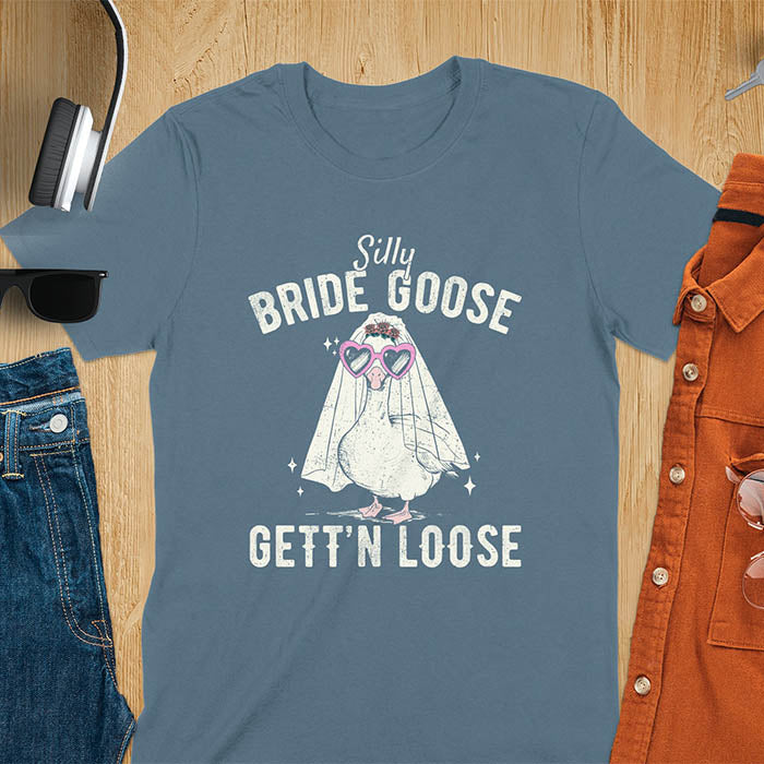 A t-shirt with a goose dressed as a bride with heart sunglasses and a veil. Text: "Silly BRIDE GOOSE" above and "GETT'N LOOSE" below. This funny bride shirt features a silly goose for getting ready bridal. A bride crewneck and engaged gift, a fiancé gift for honeymoon outfit or honeymoon gifts. A married couple shirt or new bride shirt, a fun engagement announce idea. A couple's tee-shirt and funny goose gift.